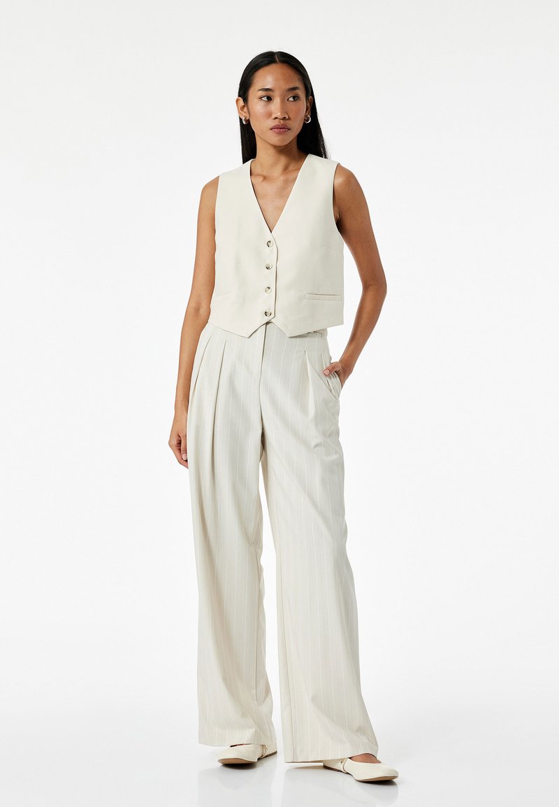 Gilet avorio corto con cinque bottoni e una tasca, abbinato a pantaloni a gamba larga color crema con righe sottili. Il materiale sembra liscio e leggero.