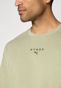 Alyvuogių žalia medvilninė marškinėlė su apvalia iškirpte, trumpomis rankovėmis ir siuvinėtu "HYROX" logotipu bei juodu "Puma" emblemą ant krūtinės.