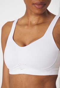 Hvit sports-BH laget av mykt stoff, med brede stropper, v-halsdesign og små ventilasjonshull nederst. Nike-logoen er synlig.