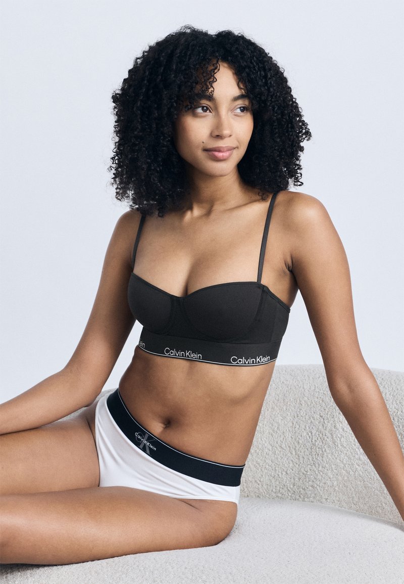 Νεαρή γυναίκα με κατσαρά μαλλιά που φορά μαύρο σουτιέν και λευκό εσώρουχο με το λογότυπο Calvin Klein, καθισμένη σε υφασμένη ανοιχτόγριζα επιφάνεια.