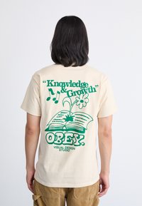 Camiseta de algodón beige con texto gráfico e imágenes en verde. El diseño incluye un libro abierto, una flor y la palabra "OBEY." en un tipo de letra en negrita.