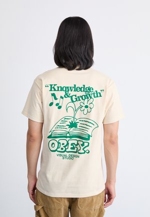 Camiseta de algodón beige con texto gráfico e imágenes en verde. El diseño incluye un libro abierto, una flor y la palabra "OBEY." en un tipo de letra en negrita.