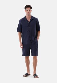 Marineblaues Kurzarmhemd mit Knöpfen und passender Shorts, aus strukturiertem Stoff. Getragen mit schwarzen Sandalen, stehend vor einem schlichten Hintergrund.