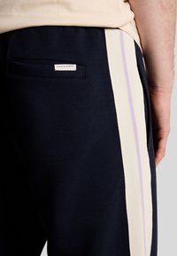 Marineblauwe trainingsbroek met crèmekleurige zijkanten en een lavendelaccent. Heeft een labelzakje op de achterkant met "Lyle & Scott" branding.