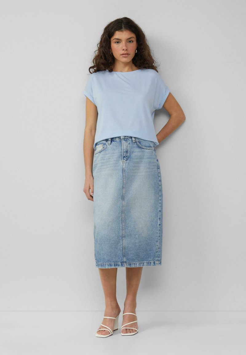 Lichtblauwe top met korte mouwen, gecombineerd met een spijkerrok tot op de knie. De outfit heeft een ontspannen pasvorm en een subtiele textuur. Duidelijke witte sandalen maken de look compleet.