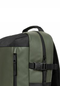 Mochila verde oliva con acentos negros, que presenta un cierre con cremallera, un bolsillo lateral, correas y detalle de hebilla. Fabricada con un material duradero y texturizado.