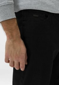 Schwarze Denim-Jeans mit Slim-Fit, ausgestattet mit einem Etikett an der Tasche und strukturierter Stoff. Eine Hand ist seitlich positioniert und hält leicht zu.