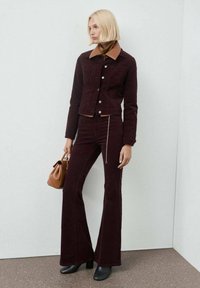 Giacca in corda bordeaux con colletto marrone, chiusura a bottoni, abbinata a pantaloni a zampa coordinati e una borsa marrone; decolleté nere eleganti.