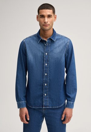 Mann trägt ein langärmliges, geknöpftes Jeanshemd und passende Jeanshose, steht vor hellem, einfarbigem Hintergrund und schaut geradeaus.