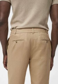 Chinos beiges avec une coupe droite, taille élastique, deux poches arrière avec fermetures à bouton, et une texture de tissu lisse.