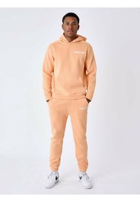 Sudadera y joggers de color melocotón, hechos de una tela suave, con un bolsillo tipo canguro y un logo impreso en blanco en el pecho y el muslo.