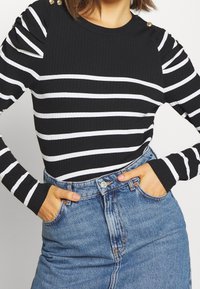 Schwarz-weiß gestreiftes, geripptes Langarmshirt mit gepufften Schultern und goldenen Knopfakzenten, kombiniert mit blauen, hochtaillierten Jeans.