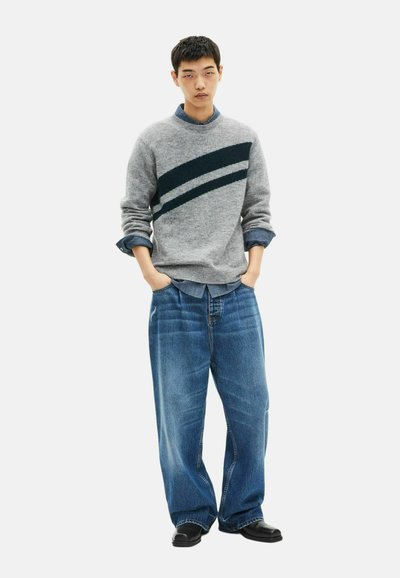 Junger Mann, der einen grauen Pullover mit dunklen diagonal gestreiften Mustern über einem blauen Hemd und weiten blauen Jeans trägt, mit Händen in den Taschen, stehend vor einem weißen Hintergrund.