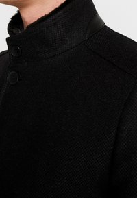 Veste en tissu noir texturé avec un col montant, quatre boutons et des accents en cuir sur le col et les coutures des épaules.