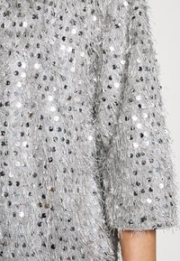 Silverfärgad paljettopp med texturerad fransade tyg, korta ärmar och lös passform. Reflekterande paljetter skapar ett glittrande mönster överallt.