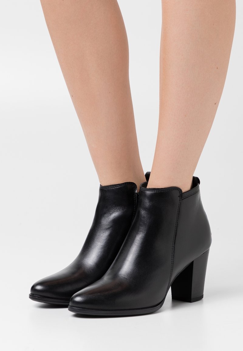 Bottes noires en cuir montant à la cheville avec talons carrés portées sur des jambes nues sur fond blanc.