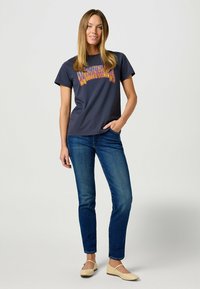 Camiseta corta de color navy con un gráfico de texto colorido, combinada con jeans ajustados azules y zapatos planos de color claro. Diseño sencillo y ajuste cómodo.
