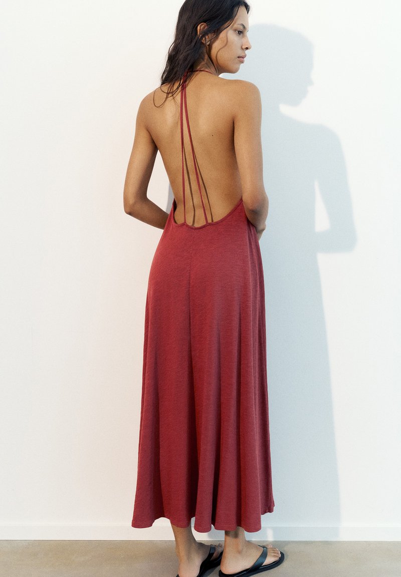 Femme portant une robe rouge sans manches et dos nu avec de fines bretelles, debout pieds nus contre un mur blanc uni.