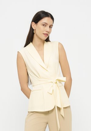 Žena s dlhými tmavými vlasmi, oblečená v krémovom bezrukávovom zavinovacom saku previazanom mašľou a béžových nohaviciach, stojaca pred bielym hladkým pozadím.