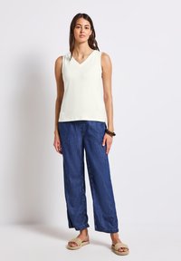 Top senza maniche di colore crema chiaro con scollatura a V, abbinato a pantaloni in denim blu scuro larghi. La modella indossa sandali slip-on color cammello.