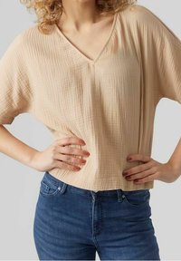 Haut beige clair texturé avec encolure en V et manches courtes, associé à un jean denim bleu. Le tissu a un aspect froissé.