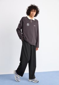 Mørkegrå langærmet polo med beige krave, logo detaljer og afslappet pasform. Bæres med vide sorte bukser og grå-blå sneakers.