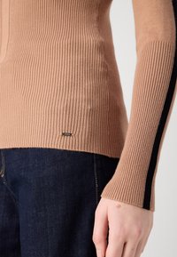 Haut à manches longues côtelé camel avec une bande noire sur la manche, porté avec un jean bleu foncé, montrant un gros plan du torse et de la main.