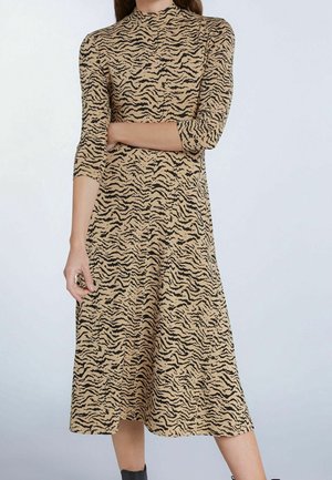 Vrouw draagt een beige-zwart midi-jurk met zebraprint en driekwart mouwen, staand tegen een lichte achtergrond.