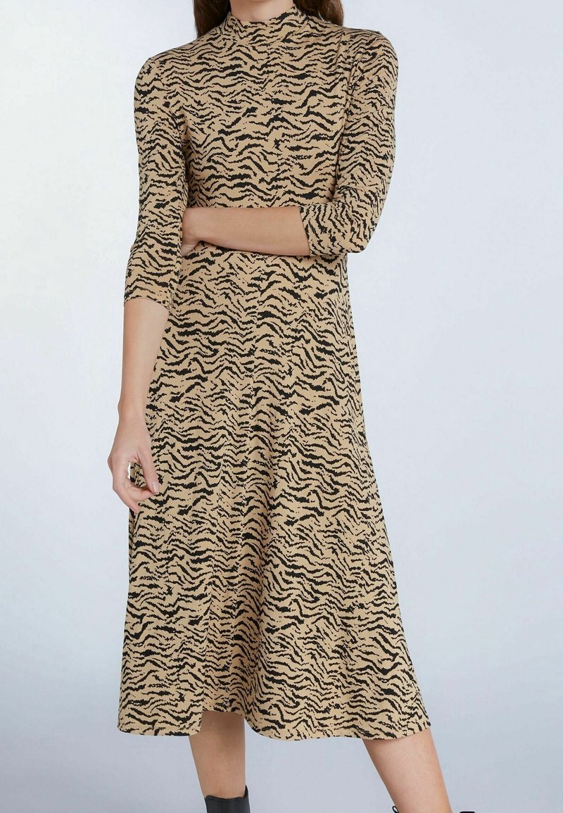 Femme portant une robe midi beige et noire à imprimé zèbre avec des manches trois-quarts, debout sur un fond clair.