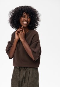 Glunderende vrouw met krullend haar draagt een bruine trui met korte mouwen en hooggetailleerde grijze broek, terwijl ze poseert met haar handen bij haar gezicht.