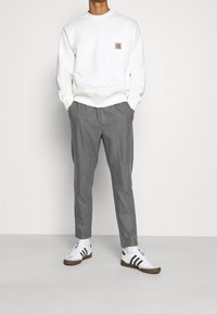Person iført en hvid sweatshirt med brystlomme, grå tapered bukser, hvide sokker og hvide sneakers med sorte striber.