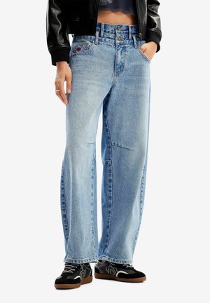Jeans de tiro alto en azul claro con un corte relajado, que cuentan con bolsillos delanteros y una cremallera. Combinados con zapatillas negras y una chaqueta de cuero.