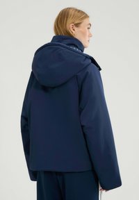Granatowa pikowana kurtka z dużym kapturem, o gładkiej fakturze i zwężonym dole. Subtelne detale ściegów na kapturze.