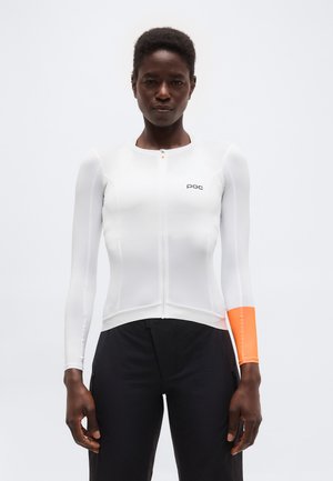 Γυναίκα που φοράει εφαρμοστή λευκή αθλητική ζακέτα με μακριά μανίκια και φερμουάρ μπροστά, πορτοκαλί λωρίδα στη μανσέτα του αριστερού μανικιού, και μαύρο παντελόνι, σε λευκό φόντο.