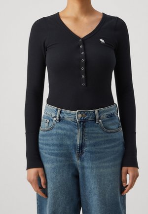 Frau trägt ein schwarzes langärmliges Henley-Oberteil mit silbernen Knöpfen und hellblaue Jeans mit hoher Taille, stehend vor einem schlichten Hintergrund.