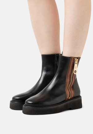 Botas de cuero negras hasta el tobillo con un cierre lateral con detalle dorado y una franja marrón. La suela es texturizada y ligeramente elevada.