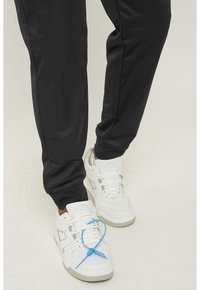 Witte sneakers met een gestructureerd ontwerp en een blauwe vetersluiting, gecombineerd met zwarte, tapered sportbroek met een gladde afwerking.