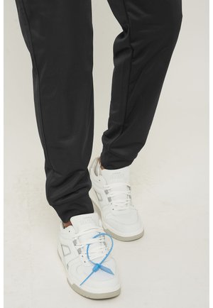 Witte sneakers met een gestructureerd ontwerp en een blauwe vetersluiting, gecombineerd met zwarte, tapered sportbroek met een gladde afwerking.