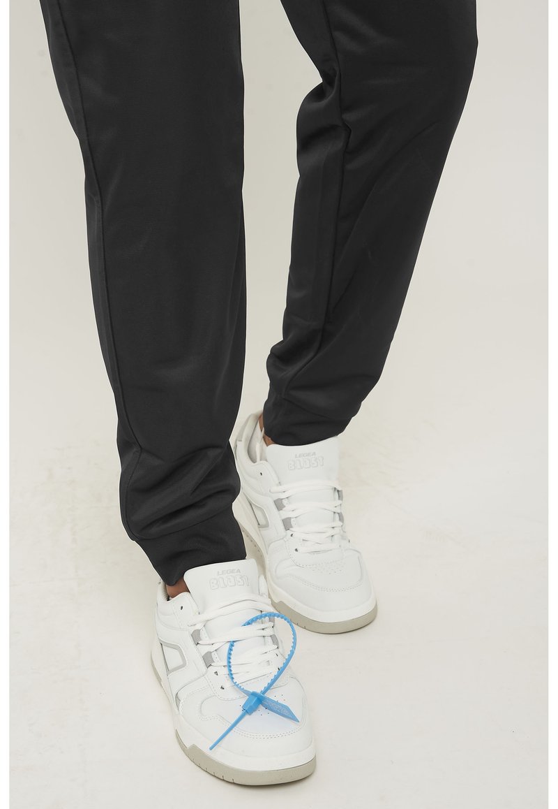 Witte sneakers met een gestructureerd ontwerp en een blauwe vetersluiting, gecombineerd met zwarte, tapered sportbroek met een gladde afwerking.