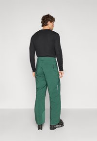 Žalios spalvos izoliuotos slidinėjimo kelnės su laisvu kirpimu, su užtrauktukais kišenėse ir logotipu. Derinamos su juoda ilgomis rankovėmis marškinėliu.