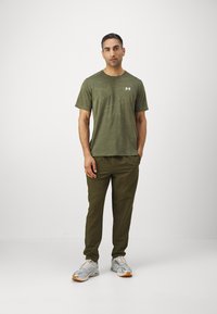 Grøn camouflage-musteret kortærmet skjorte med rund hals, parret med olivengrønne tapered bukser og grå sneakers med orange detaljer.