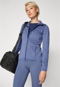 Blå idrettsjakke med glidelås, teksturerte ermer og sidelommer; kombinert med matchende leggings og en svart veske med nett- detaljer.
