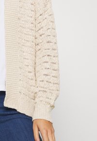 Ljusbeige öppen stickad cardigan med ribbade kanter. Den har långa ärmar och en lös passform. Bärs över en vit topp och matchas med mörka jeans.