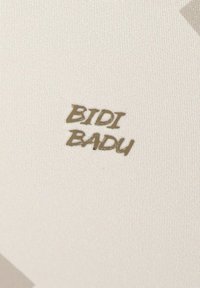 Het label toont de tekst "BIDI BADU" in een gestileerd, metallic gouden lettertype op een gestructureerde off-white stoffen achtergrond.