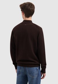 Junge Person, die mit dem Rücken zur Kamera steht, trägt einen dunkelbraunen Pullover und eine blaue Jeans vor einem hellen einfarbigen Hintergrund.