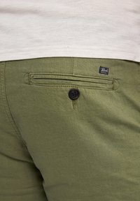 Olive green shorts with a textured fabric, featuring a back pocket secured by a dark, circular button and a branded tag on the waistband.