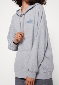 Sudadera de algodón gris con un bolsillo frontal, capucha con cordones y logo "silverTab" en azul y rosa. Presenta puños y dobladillo acanalados.