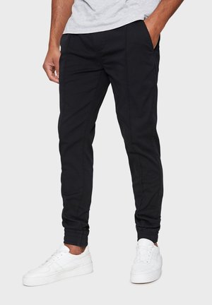 Homme portant un pantalon de jogging noir et des baskets blanches, debout avec une main dans la poche contre un fond clair uni.