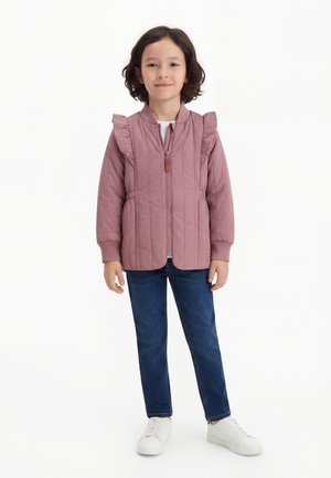 Barn med mørkt hår iført en støvlilla vattert jakke med volanger, blå jeans og hvite joggesko, stående mot en hvit bakgrunn.