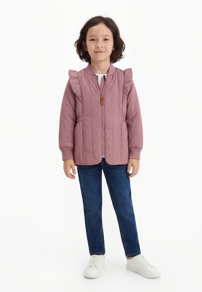 Παιδί με σκούρα μαλλιά που φοράει μια μωβ καπιτονέ ζακέτα με βολάν, μπλε τζιν και άσπρα αθλητικά παπούτσια, στέκεται μπροστά σε λευκό φόντο.